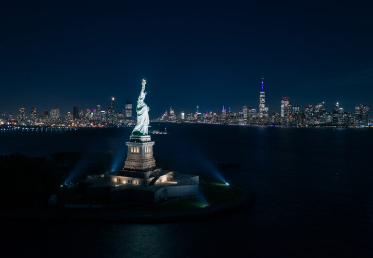 Aerial view of the Statue of Liberty at night with the Manhattan skyline, by Arte City Media