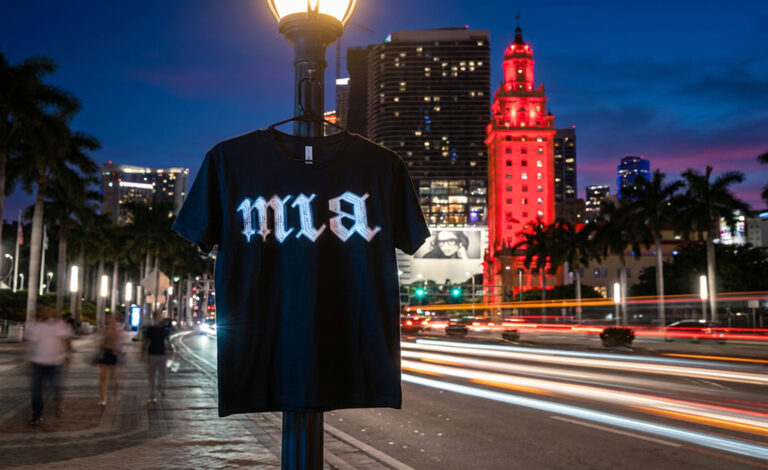 Black MIA blackletter t-shirt hanging on a street lamp in downtown Miami at night with long-exposure light trails, product photography by Arte City Media
